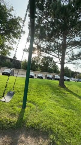 The video shows a swing set with a single swing hanging from it. The swing is empty. The swing set is located in a grassy area with trees and a building in the background. Several cars are parked in a lot behind the building. The sun is shining through the trees, creating lens flare. The camera is positioned to show the swing and its movement.