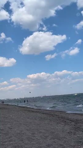 The video shows a view of a beach with a body of water. The sky is blue with scattered white clouds. The beach is made of dark grey pebbles or small rocks. There are a few people visible along the shoreline, some walking and others in the water. The water has waves and appears to be a sea or large lake. In the distance, there are some structures or buildings along the coastline and a few boats or other watercraft on the water. There is also a dark object, possibly a kite or a drone, visible in the air above the water. The overall scene depicts a sunny day at the beach.