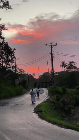 The video shows a rural street at sunset. The sky is a mix of pinks and oranges. A woman in a white dress walks away from the camera on the wet asphalt, followed by a person riding a bicycle.  Utility poles with wires line the road.  Vegetation grows thick on either side, including palm trees.  A railroad crossing is visible further down the road.