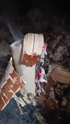 A close-up video shows a toilet paper roll igniting in a fireplace. The roll is placed amongst burning logs and embers, with flames and smoke rising from it. The outer layers of the roll are burning, revealing the cardboard interior. The surrounding environment appears to be dark and smoky.