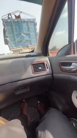 The video shows the interior of a car, specifically the passenger side. The dashboard is visible with an air vent featuring a wood-style trim. Below, a glimpse of someone's feet, with painted white toenails, rests on the floor mat. The most prominent feature of the video is the cracked windshield, through which a large blue truck can be seen moving alongside the car. The view through the side window shows the truck passing, indicating the car is also in motion. The environment appears to be a road or highway with trees visible in the distance.