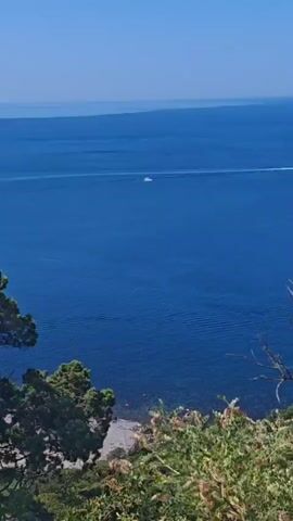 The video begins with a wide shot of a calm, blue ocean under a clear sky, with a small sailboat visible in the distance. Trees in the foreground partially obscure the view. The camera then pans slightly to reveal a pier extending into the water with lights on it. The scene shifts to a panoramic view of a coastal town nestled among green hills. The camera then focuses on a woman walking away from the camera on a dirt path. She is wearing a white shirt and light blue shorts and sandals. The background shows a lush green hillside with the ocean and the town visible in the distance. The woman stops and adjusts her hair, looking out at the view. The video ends with a wider shot of the woman on the path with the scenic landscape behind her.