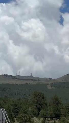The video shows a mountain range under a partly cloudy sky. The foreground features a dense forest of green pine trees. The middle ground shows a sloping, grassy mountain covered with smaller scattered vegetation. The background reveals a larger, rocky mountain peak partially obscured by white clouds. The sky transitions from a bright blue with scattered white clouds to a more overcast scene with larger, darker clouds. The camera appears to be stationary, capturing a panoramic view of the landscape.