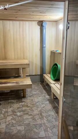 The video shows the interior of a sauna. Wooden benches are visible, arranged in a tiered configuration. A grey pipe runs vertically along the wall. Two green buckets and a yellow ladle are placed on one of the benches. The floor has a tiled pattern. The walls and ceiling are paneled with wood. The overall lighting is dim. There are no people or animals present in the video.