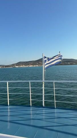 The video is a vertical shot from the deck of a boat. A white railing with vertical supports runs horizontally across the frame. A white flagpole stands in the center, with the Greek flag flying from it, its blue and white stripes rippling in the wind. The sea is a deep blue-green, with small waves visible. In the background, a coastline is visible, with hills covered in vegetation and a town with white buildings clustered along the shore. The sky is a clear, bright blue. The sun appears to be low in the sky, casting long shadows on the blue deck of the boat. The camera is stationary.