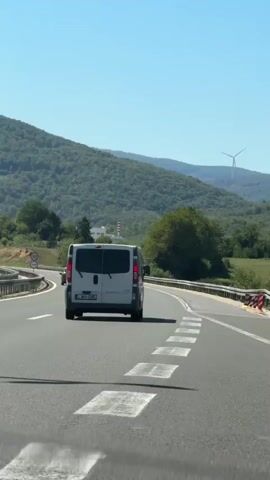 The video shows a view from inside a vehicle traveling on a highway. In front of the camera is the back of a white van. The highway has multiple lanes, and there are other cars on the road, including two dark-colored cars ahead of the van. Speed limit signs indicating 80 are visible on both sides of the road. The highway curves to the left, with a guardrail along the side. Beyond the highway, the landscape is dominated by a large, tree-covered hill under a clear blue sky. In the distance, on the right side of the frame, a wind turbine can be seen. The overall scene suggests a sunny day in a rural or mountainous area.