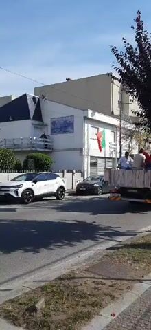 A slow-motion video captures a street scene with a white car parked on the left side of the road. A dark-colored car is parked behind it. A flatbed truck, filled with people, drives down the street. A Portuguese flag is displayed prominently on the side of a building. A person stands on the sidewalk, facing the truck. Another person is also visible on the sidewalk. Trees line the side of the road. The sky is clear and blue.