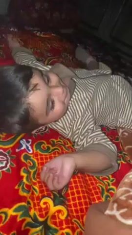 A young child with dark hair and a striped outfit lies on a bed with a vibrant red and gold patterned blanket. They initially smile and giggle, then become more subdued, looking off to the side. A person's hand and part of their arm are visible in the bottom right corner of the frame. A blue object, possibly a toy or part of the bedding, is seen momentarily. The environment appears to be indoors, possibly a bedroom.