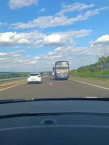 A first-person view from a vehicle on a highway shows a white car in front, followed by a black double-decker bus. The bus has the logo "RENTALTRAVEL" on its side. The sky is blue with scattered white clouds. The highway has multiple lanes and a grassy shoulder bordered by a metal guardrail. The landscape on the left appears to be open fields or water, and the right side has trees and hills. The video captures the vehicles traveling at speed on the highway under a clear sky.