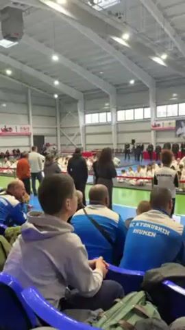 The video shows a large gymnasium with a karate tournament underway. A crowd of spectators are seated on blue chairs, many wearing blue jackets with "FC KYOKUSHIN SHUMEN" written on them. The background features a green and blue sports court where a large group of participants in white karate uniforms are seated and standing. Several adults, likely coaches or officials, are standing in front of the group. The gymnasium has a high, metallic roof with bright overhead lighting. Several large posters are displayed on the walls. People are moving around, and a woman in black appears to be addressing the group. The atmosphere seems to be focused and attentive.