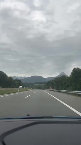 This is a first-person perspective video of driving on a highway. The road has multiple lanes with white lines marking the boundaries. The sky is overcast and cloudy. On the left side of the road, there are trees and some distant hills or mountains. On the right side, there is a metal guardrail and more trees. A few vehicles are visible in the distance on the highway. The dashboard of the car is partially visible in the foreground.