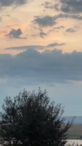 The video displays a time-lapse of a sunset. The sky is filled with dynamic clouds, showcasing a blend of grays and oranges. In the foreground, a dark-leafed tree stands prominently. Below the tree, a body of water is visible, with hints of a landscape and fields stretching into the distance. The overall scene evokes a peaceful, natural setting.