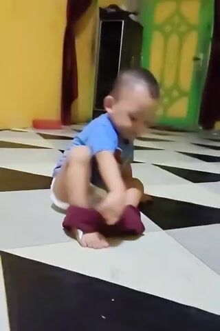 A child wearing a blue t-shirt and a diaper is sitting on a black and white tiled floor. The child is pulling a maroon garment down over their right foot. The background shows a yellow wall with a dark red curtain on the left and a dark cabinet on the right. A green door with a decorative panel is visible in the background.