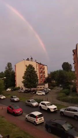 This is a video of a rainbow appearing over a city street. The scene is set in Częstochowa, Poland, and shows a residential building with balconies, trees, and several parked cars. A rainbow arches across the cloudy sky, which suggests it might be a rainy or post-rainy day. The time of day appears to be late afternoon or early evening, given the muted light. The overall mood is serene, with the natural phenomenon of the rainbow adding a touch of wonder to the urban landscape. No people are visible in the video, but the presence of parked cars indicates a residential area. The video is of good quality, capturing the colors of the rainbow clearly against the subdued sky.