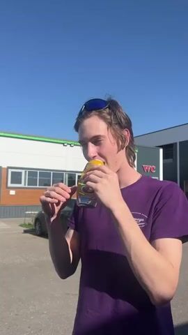 A young man with long brown hair, wearing a purple t-shirt and sunglasses, is standing outdoors, holding a yellow and green beverage can to his mouth. He is making a loud, sustained, drawn-out vocalization, possibly a scream. The background shows a gray car, a building with green trim and several windows, and a clear blue sky.