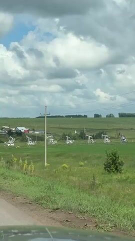 The video shows a wide, green, rural landscape under a cloudy sky. In the foreground, a dirt road is visible at the bottom. Several white and red pumpjacks, used for oil extraction, are scattered across a large, grassy field. In the middle ground, a tall, gray utility pole stands with wires running across the frame. A road sign with the number 75 is visible on a pole to the left of the utility pole. In the background, a line of trees and rolling hills stretch across the horizon. The pumpjacks appear to be operational, with their arms moving up and down. The overall impression is one of industrial activity in a natural setting.