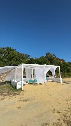 The video shows a white, open-air cabana on a sandy beach. The cabana has a white roof and white curtains that are blowing in the wind. Inside the cabana are two teal lounge chairs. Behind the cabana, there is a lush green hillside covered in trees. The sky is clear and blue. The beach appears to be empty of people.