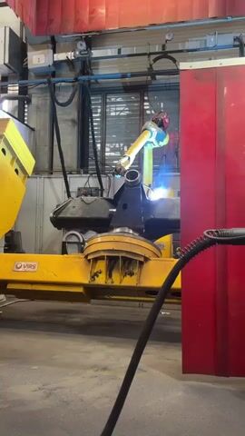 A robotic arm is welding metal in a factory setting. The robot is yellow and black and is positioned on a yellow base. It has a welding torch attached to its arm, which is producing a bright blue light as it welds. The surrounding environment is industrial, with metal walls, red curtains, and visible pipes and wiring. A black hose extends from the bottom right of the frame towards the welding area.