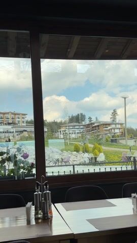 The video presents a view from inside a restaurant, looking out through a large window. Tables with black chairs and set places are visible. Condiment containers stand on the tables. Outside, there is an artificial pond with rocks and landscaping. In the background are several large, multi-story buildings that appear to be part of a resort or hotel complex. The sky is cloudy with patches of blue visible. There is no one visible in the video frame.