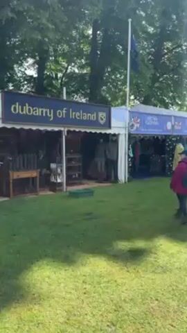 Here is a concise description of the video:

The video shows a Dubarry of Ireland booth at an outdoor event. The booth is set up under a white canopy with the company's sign displayed prominently. The booth is filled with clothing, shoes, and boots. There are mannequins dressed in Dubarry apparel and a man sitting inside the booth. Outside the booth, a person with a backpack walks by. The scene is set on a grassy field, with trees and other booths visible in the background.