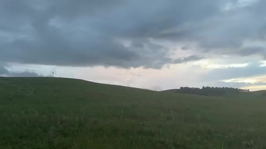 A wide shot captures a rolling green hill under a cloudy sky. A small, dark object resembling a weather vane or sculpture stands on the crest of the hill to the left, silhouetted against the brighter sky. In the distance to the right, a dark line of trees borders the horizon. The grass in the foreground is textured and appears windblown. The lighting suggests either dawn or dusk, with diffused light filtering through the clouds. The camera pans slightly to the right, revealing a slight dip in the landscape before the ground rises again towards the treeline. No people or animals are visible in the frame.