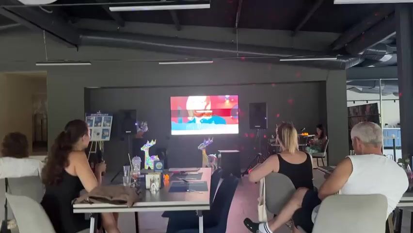 This media file captures an indoor gathering, possibly a party or a presentation, with a group of people seated at tables facing a large screen. The room appears to be a modern, dimly lit space with exposed pipes and industrial-style lighting on the ceiling. Disco lights are projecting colorful spots onto the ceiling and walls, suggesting a festive atmosphere.

The central focus is the large screen displaying a video feed of a person, likely giving a presentation or performing. Two speakers are positioned on either side of the screen. In the foreground, three individuals are seated at a table. A woman on the left, with long dark hair tied in a ponytail, is holding a microphone and appears to be speaking or singing. She is wearing a black dress. In the center, a woman with blonde hair, also in a black dress, is looking towards the screen. On the right, a man with gray hair and a white sleeveless shirt is seated. Another person, partially visible, is seated further back on the left. In the background, a woman is seated at a table to the right of the screen, engaged with her device.

Several decorative elements, including illuminated reindeer figures, are placed on the tables. A whiteboard or display board with some text and images is visible on the left. The overall mood seems relaxed and engaged, with the disco lights adding a playful element to the scene. The time of day is indeterminate due to the indoor setting, but the lighting suggests it could be evening. The location is identified as Даниловка (алтайский), Russia. The visible text on the display board is "ИНФОРМАЦИЯ" (Information) in Russian, with accompanying icons.