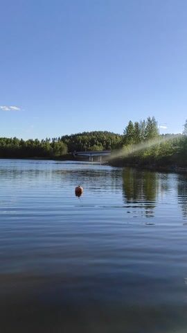 This media file depicts a scenic view of a body of water, likely a lake, bordered by a dense forest on the left. The sky is a vibrant blue with scattered white clouds, suggesting a clear and pleasant day. The time of day appears to be late morning or early afternoon, given the bright sunlight casting reflections on the water's surface.

There are several buoys floating on the water, indicating a designated area, possibly for swimming, boating, or a navigational channel. The water is calm, with gentle ripples suggesting a light breeze. In the distance, a small structure, possibly a jetty or a small building, is visible among the trees.

The overall mood is tranquil and serene, evoking a sense of peace and natural beauty. The lush green of the forest contrasts with the deep blue of the water and sky, creating a visually appealing landscape. There is no visible text in the media file. The scene is characteristic of a Finnish summer, given the location context of Joutsa, Finland, a region known for its numerous lakes and forests.