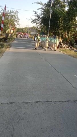 A group of approximately eight individuals dressed in light green uniforms and headscarves are walking in a single file line on an asphalt road. They appear to be marching or participating in a parade, with some of them raising their arms. The road is lined with trees and vegetation on the left side. Further down the road, several motorcycles are visible, and more people are gathered on the side. The sky is clear and bright, suggesting daytime. The camera is positioned at a low angle, looking across the road. The video is oriented vertically.