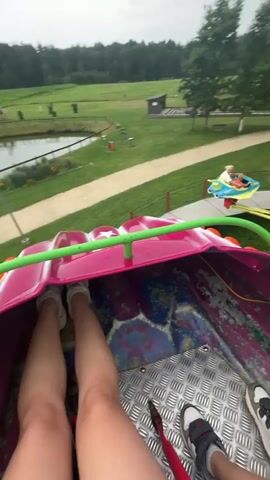 The video shows a point-of-view shot from a carnival ride. The ride appears to be a spinning amusement ride with multiple pods for passengers. The pods are colorful, with yellow and pink accents. In the foreground, we see parts of the ride the viewer is on, including a pink and purple section with some wear and tear, and a metal floor with tread. A person's legs are visible in the foreground, wearing white sneakers and dark shorts.

In the background, we see the amusement park environment. There's a grassy area with trees and a winding path. Other carnival rides are visible, including a carousel-like structure and other spinning rides with passengers. There are people walking around in the background, some standing near a red umbrella. The sky is overcast. The ride the viewer is on is actively spinning and moving.