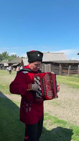 A man in a traditional red Cossack uniform with a black fur hat plays a red accordion. He stands in an outdoor setting with a bright blue sky above. Behind him are wooden buildings and some people walking in the distance. The man is focused on playing the accordion.