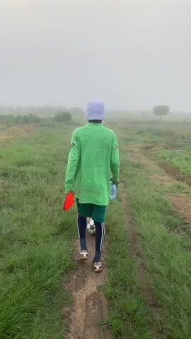This video shows a person walking away from the camera on a dirt path through a grassy field. The person is wearing a green long-sleeved shirt, green shorts, black leggings with white stripes, a light blue cap, and athletic shoes. They are carrying a soccer ball, a red cone, and a blue water bottle. The field is covered in green grass, and there are some trees visible in the distance. The sky is overcast and hazy, suggesting it might be early morning or late afternoon, or possibly foggy or rainy weather. The overall atmosphere appears calm and perhaps a bit isolated. There is no visible text in the video. The location is identified as Gwafan, Nigeria. The activity is simply walking, likely for exercise or to a practice/game.
