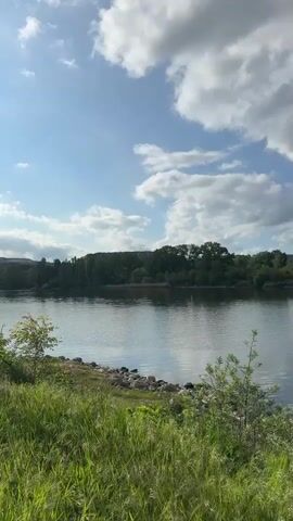 The video shows a wide river on a partly sunny day. The sky is blue with white, fluffy clouds. The shoreline in the foreground has grass and rocks. In the distance, a line of trees forms the opposite bank. A red buoy is visible in the water. Towards the end of the video, a few boats are seen docked on the far shore. A white pole is also visible at the right edge of the frame.