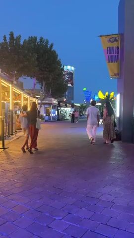 The video shows a paved walkway at dusk. Several people are walking away from the camera. On the left, two people, one wearing a blue t-shirt and khaki shorts and the other a dark top and long red skirt, walk together. Further down the walkway, on the right, two more people, one in a white shirt and white pants and the other in a patterned dress, walk side-by-side. In the background, there are various shops and stalls, some illuminated with warm lighting. A large billboard with an image of a superhero is visible atop a tall building in the distance. The sky is a deep blue, indicative of twilight. Colorful lights, some purple and pink, illuminate the pavement. A yellow banner with the words "FUN IS NOW" hangs from a pole on the right side of the frame.