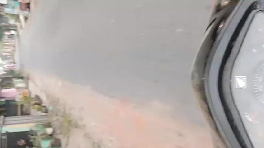 The video shows a street scene from a moving vehicle, likely a motorcycle or bicycle, due to the perspective and the vibration of the camera. The road is paved and curves to the right. On the left side of the road, there are houses and trees. Some houses have signs and flags, possibly indicating a celebration or national holiday. There are also some small shops or stalls along the roadside. On the right side of the road, there is a dirt shoulder. The sky is overcast and bright. In the distance, there appear to be more houses and trees. There are a few objects on the road, including what looks like a discarded piece of green vegetation on the right side. The video is shaky, suggesting the motion of the vehicle. There are no people clearly visible or interacting in the frame.