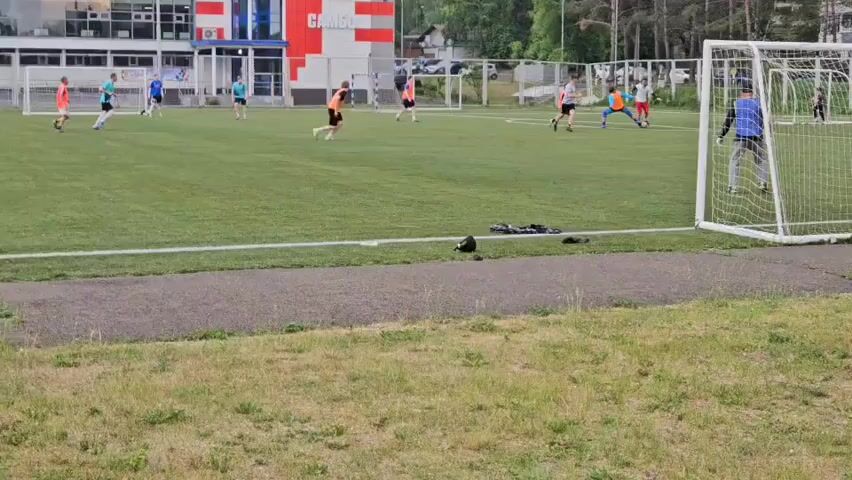 The video shows a children's soccer game on an artificial turf field. Several children in brightly colored uniforms are playing. Some are running with the ball, while others are positioned defensively. A goalkeeper is visible in front of a net on the right side of the frame. In the background, there is a building with a red and white facade and trees. The field is bordered by a running track.