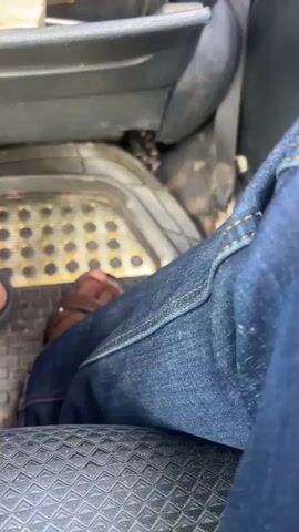 The video shows the interior of a vehicle from a low angle, focusing on the driver's feet and the floor pedals. Jeans and brown sandals are visible. The floor mats are dirty and textured. The car appears to be in motion, indicated by the blur of the exterior landscape in the later frames, which shows a bright blue sky with scattered clouds and green, hilly terrain.