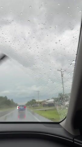 The video shows a view from inside a car through a rain-streaked windshield. Outside, it is raining, and the sky is overcast with dark clouds. A two-lane road is visible, with a car in front of the camera vehicle driving in the same direction. Other vehicles are parked or driving on the side of the road. The windshield wipers are active and sweep across the lower portion of the windshield, clearing some of the raindrops. The overall scene conveys a sense of a rainy day journey.