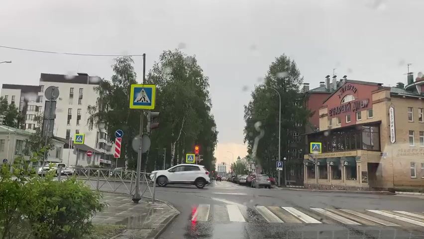 The media file shows a street scene on a rainy day in Khanty-Mansiysk, Russia. The camera is positioned inside a vehicle, looking out through a wet windshield, indicated by visible water droplets.

The foreground features a wet asphalt road with a pedestrian crossing marked by white stripes. A metal barrier separates the sidewalk from the road. To the left, there is lush green foliage.

The mid-ground is dominated by a street intersection. Traffic lights are visible, with one showing a red light for vehicular traffic and a green pedestrian signal. Several road signs are present, including pedestrian crossing signs (blue squares with white figures walking) and a circular red and white sign, likely a "no entry" or "no stopping" sign. Tall, leafy trees line the sides of the street, their leaves appearing dark and wet. Cars are visible on the road, stopped at the intersection.

In the background, buildings line the street. On the right, a brick building with arched windows and a sign reads "СТАРЫЙ ДВОР" (Stary Dvor), which translates to "Old Courtyard." This building appears to house a restaurant or cafe, with outdoor seating visible. Further down the street, more buildings and trees can be seen. The sky is overcast and gray, consistent with the rainy weather. The overall impression is a quiet, somewhat somber street scene during a rain shower. No people are clearly identifiable. The time of day appears to be daytime, given the ambient light, though the overcast sky makes it difficult to determine precisely.