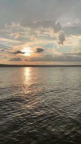A video clip shows a wide expanse of water, likely a lake or ocean, under a cloudy sky. The sun is setting on the left side of the frame, casting a golden reflection on the water. In the distance, a thin line of land is visible, and on the right side of the frame, a small wooden dock or pier extends into the water, with a cluster of green foliage growing around it. The water is gently rippling, suggesting a calm day. There are no people or animals visible in the video.
