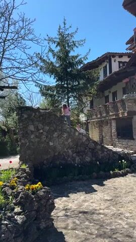 Two children are playing on a stone structure in front of a building on a sunny day. The building has a traditional design with a balcony. One child is standing on the top of the structure, while the other is climbing up the side. Flowers are planted around the base of the structure.