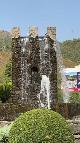 The media file shows a stone fountain designed to resemble a castle turret. Water cascades down the sides and sprays from the bottom, creating a dynamic display. In front of the fountain is a neatly trimmed green bush. Behind the fountain, a hillside with green vegetation and some buildings is visible under a clear blue sky, suggesting a warm, sunny day. A billboard advertisement for "FUENTE LARIOS" apartment sales is prominently displayed to the right of the fountain. The text on the billboard reads "New 2 & 3 bed apartments" and "Sales Office in Benahavís centre." A phone number, "900 13 00 44," is also visible. In the background, power lines and pylons are visible on the distant hills, and a street lamp and palm trees are also part of the scene. The overall impression is one of a well-maintained public space in a sunny, likely Mediterranean, location. There are no people visible in the media file.