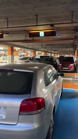A view from inside a parking garage shows a line of cars parked in designated spots. Overhead lights illuminate the concrete ceiling and the cars. Signs with accessibility symbols are visible above some of the parking spaces. The camera is positioned low, looking towards the rear of a silver Audi A3 in the foreground, with its license plate partially visible. Other vehicles, including a white Tesla, a black SUV, and a red car, are parked further down the aisle. The parking lot has orange and blue markings on the floor.