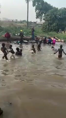 The video shows a group of children playing in a river or stream. The water appears somewhat murky or brownish. The children, mostly undressed, are splashing, swimming, and interacting with each other in the water. On the banks of the river, there are other individuals, some who appear to be adults, near piles of what looks like clothing or laundry. The environment is somewhat rural, with trees and some structures visible in the background. The overall scene suggests a community activity involving both recreation and domestic tasks taking place near a water source.