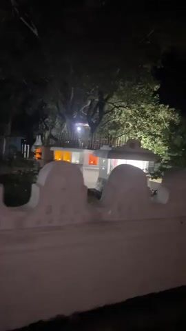 The video pans from left to right at night, showing a white, low-lying wall in the foreground with a decorative, undulating top. Beyond the wall, a white building is visible, illuminated by lights. Orange and yellow lights are visible inside or on the building. Trees with dense foliage are present behind and around the building, with a bright spotlight illuminating some of the branches and leaves. To the right of the building, a row of standing yellow statues, possibly of monks, are lined up on a platform. Palm trees are visible in the background on the right side of the frame. The scene appears to be outdoors and illuminated artificially.
