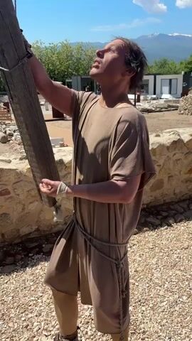 The video shows a man dressed in a toga and rope belt standing outside on a bright day. He is holding a wooden pole and looking upwards towards the sky. The camera follows his gaze upwards to a wooden crane structure with a rope extending down. The sky is blue with scattered clouds and mountains are visible in the distance. The man then looks down, suggesting he is overseeing construction work. The ground is covered with gravel and there are stone walls nearby.