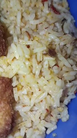 A close-up shot of a plate of white rice. The rice appears to be seasoned with red chili flakes and possibly other spices, giving it a slightly yellowish tint. There are also some darker specks mixed into the rice, which could be small pieces of meat or vegetables. On the left side of the frame, a portion of a golden-brown, fried food item is visible, partially obscured by the rice. The plate is a bright blue color, suggesting it is likely made of plastic. The lighting is somewhat harsh, creating highlights on the rice grains.