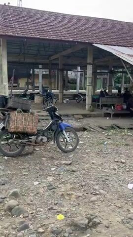 The video shows a street scene in what appears to be a small town or village. There are several buildings lining the street, some of which are shops. In the foreground, two motorcycles are parked on a gravelly surface. One is a red scooter, and the other is a black motorcycle with a person sitting on it. In the background, there are people walking and standing around, and some are interacting with each other. There are also trees and other vegetation visible. The overall impression is one of everyday life in a relatively simple setting.