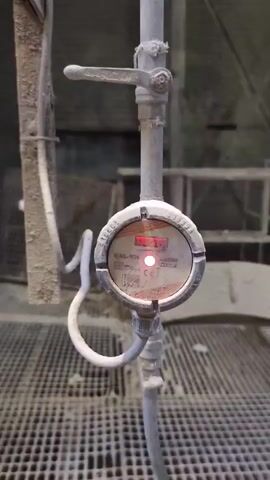The video shows a close-up of a circular gauge attached to a pipe. The gauge has a digital display that shows red numbers, and a small red light is illuminated. The gauge is dusty, suggesting an industrial or outdoor environment. A valve is visible on the pipe above the gauge. The background is out of focus but appears to be a grid-like structure and possibly some machinery or scaffolding. The overall impression is one of an industrial setting, possibly related to measurement or monitoring of some process. The text visible on the gauge includes "MAL-KH", "CE", and some other markings that are difficult to read due to the dust and resolution. The video appears to be taken in daylight, but the lighting is not strong enough to determine the time of day precisely. There are no people visible in the video. The activity depicted is simply the gauge displaying readings, with no other actions taking place.