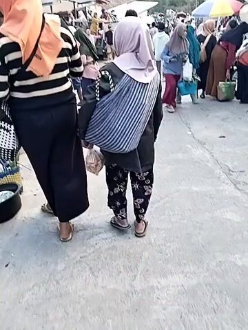 The video shows a crowded outdoor market with many people walking along a paved road. On the left side of the road, there are buildings that appear to be shops or stalls, with merchandise displayed outside. On the right side, there are more stalls and people browsing. The market is situated in a hilly, rural environment, with trees and mountains visible in the background. The sky is bright, suggesting it is daytime. People are wearing casual clothing, with many women wearing headscarves. Some individuals are carrying bags or baskets, indicating they are shopping. There is a general sense of activity and movement throughout the scene. The perspective is from someone walking within the market, looking forward at the crowd and the stalls.