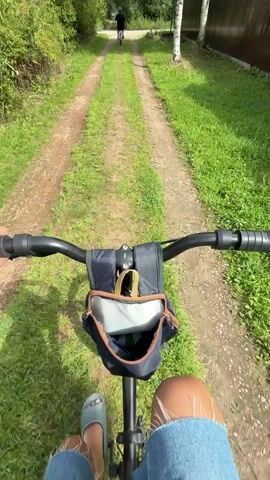 A first-person perspective video shows someone riding a bicycle on a dirt road. The handlebars of the bicycle are visible in the foreground, with a small bag attached to them. The road is flanked by green grass and trees. In the distance, another person on a bicycle is visible, riding away from the camera. The sky is cloudy.